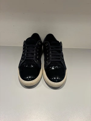 Svarta sneakers från Lanvin i mocka - Snygga svarta sneakers från Lanvin med ovandel i mocka och lackad tå. Skorna har vita sulor, svarta skosnören och metallfärgade öljetter. Perfekta för dig som gillar stilrena och exklusiva sneakers med en twist. Box fås med!!