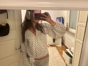 Vit prickig omlottblus med knyt - Säljer en vit blus med svarta prickar och omlottmodell. Blusen har ballongärmar med resår vid ärmslut och ett snyggt knyt framtill i midjan. V-ringad hals och luftig passform, perfekt för dig som gillar trendiga detaljer och mönster.