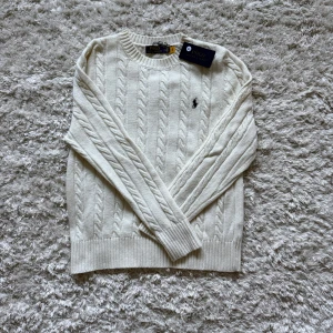 Vit kabelstickad tröja Polo Ralph Lauren - Snygg vit kabelstickad tröja från Polo Ralph Lauren med klassisk rund hals och broderad logga på bröstet. Tröjan har långa ärmar och ribbade muddar vid ärmslut och nederkant. Perfekt för dig som gillar stilrena och tidlösa plagg. Helt nyskick endast testad. Nypris 2400 mitt pris 1000