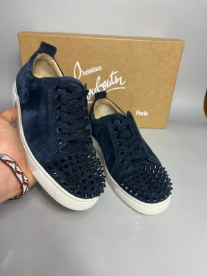 Mörkblå Christian Louboutin sneakers - Snygga mörkblå sneakers från Christian Louboutin i mocka med vita sulor och ikonisk röd undersida. Tåpartiet är täckt av svarta nitar som ger en edgy vibe. Skorna har rund tå, snörning och lyxig känsla, perfekt för dig som gillar statement pieces.