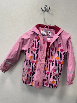 Rosa regnjacka med nagellacksmönster - Säljer en rosa regnjacka från Lupilu med coolt nagellacksmönster i lila, orange och mörkrosa. Jackan har huva med röd fleecefoder, reflexdetaljer på ärmar och nederkant samt dragkedja framtill. Perfekt för dig som gillar färg och unika prints!