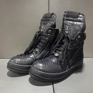 Svarta boots med reptilmönster och snörning - Unika svarta boots från Chrome Hearts med reptilmönstrad skinnyta, högt skaft och snörning. Dekorerade med metallkors på plösen och detaljer i silver. Grov platt sula och dragkedja på sidan för enkel påtagning. Perfekt för dig som gillar edgy och exklusiv stil.
