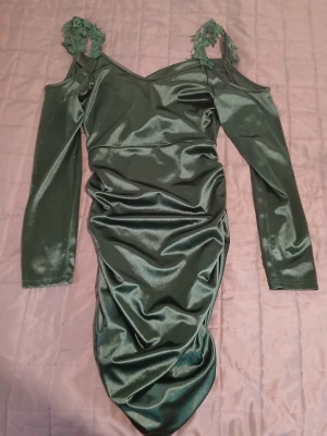 Grön långärmad klänning från SHEIN - Snygg grön klänning från SHEIN i storlek M med glansigt tyg och långärmad design. Axelbanden har fina spetsdetaljer med blommor och offshoulder-stil. Klänningen är figurnära och har en draperad effekt längs kjolen för extra stilpoäng.