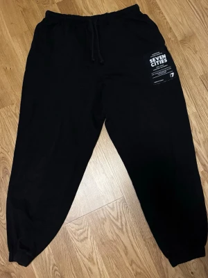 Svarta mjukisbyxor Divided H&M M - Svarta mjukisbyxor från Divided H&M i storlek M. Byxorna har resår och snörning i midjan, muddar vid bensluten och en vit tryckt detalj med texten 'Seven Cities' på ena benet. 