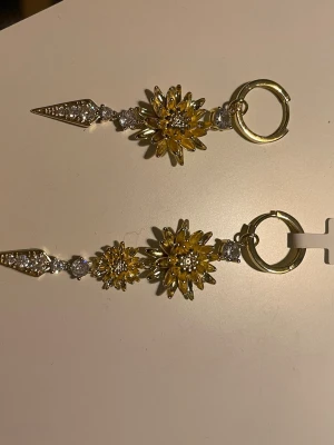 Guldiga örhängen från Ioaku - Säljer ett par riktigt snygga örhängen från Ioaku i guldton med blommiga detaljer och gnistrande stenar. Örhängena har en cool spetsig avslutning och hängs på örat med en rund ring. .