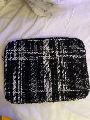Rutig datorväska från H&M - Säljer en svart och vit rutig smink väska från H&M med grovt vävt tyg och dragkedja upptill. Perfekt för att bära din laptop eller surfplatta. Stilen är klassisk men ändå trendig med sitt rutiga mönster och mjuka material.