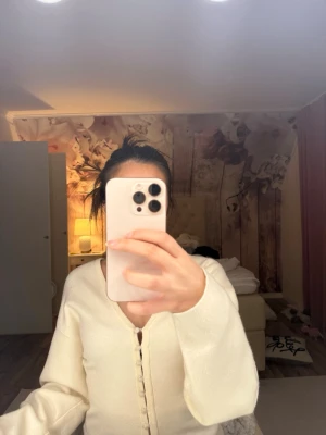 Krämvit kofta med knappar - Säljer en krämvit kofta med v-ringning och dekorativa knappar framtill. Koftan har långa ärmar och är gjord i ett mjukt material som känns skönt mot huden. Perfekt att slänga på sig när det är lite kyligare ute. I storlek s men passar mig som e xs