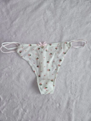 Vit stringtrosa med jordgubbsmönster - Supersöt vit stringtrosa med små rosa jordgubbar och blommor, dubbla tunna sidoband och en liten rosett med hjärtberlock framtill. Materialet är mjukt och stretchigt med meshkänsla, perfekt för dig som gillar gulliga detaljer. Helt nya utan tags. Står storlek L men uppskattar att dom passar XS-S. 