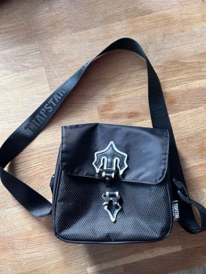 Svart axelväska från Trapstar - Säljer en svart axelväska från Trapstar med justerbar rem och coola metallspännen framtill. Väskan har ett mönstrat material på framsidan och dragkedja upptill. 