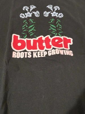 Mörkgrön Butter t-shirt  - Svart t-shirt från Butter med färgglatt tryck på bröstet. Trycket har texten 'Roots Keep Growing' och motiv med blommor och gröna blad. T-shirten har rund hals och korta ärmar, tillverkad i mjuk bomull. Perfekt för dig som gillar streetwear och grafiska prints.