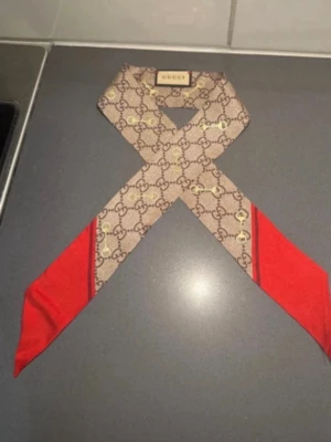 Gucci scarf med GG-mönster och rött - Säljer en lyxig scarf från Gucci med klassiskt GG-mönster i beige och brunt, detaljerad med guldfärgade hästbitsmotiv och röda ändar. Scarfen är smal och lång, perfekt att knyta runt halsen eller i håret för en trendig touch. Kommer med originalförpackning och tagg.