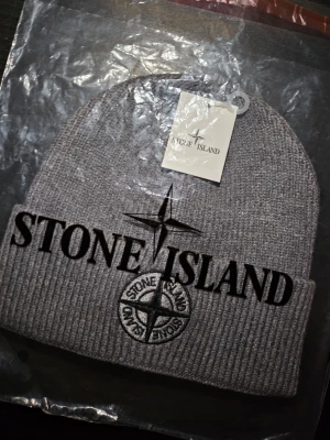 Stone Island mössa - Säljer en grå mössa från Stone Island med klassisk logotyp framtill. Mössan är ribbstickad och har uppvikt kant för extra komfort. Perfekt för att hålla värmen och samtidigt se snygg ut. MED PRISLAPP, HELT NY! Jag finns även på vinted (kevorkkihiayan). Mössan passar både mån och kvinnor. 