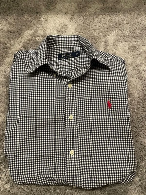 Rutig skjorta från Polo Ralph Lauren - Säljer en klassisk rutig skjorta från Polo Ralph Lauren i marinblått och vitt. Skjortan har lång ärm, knappar framtill och den ikoniska röda loggan broderad på bröstet. Tillverkad i mjuk bomull och passar perfekt till jeans eller chinos. Jag är 178 cm lång och väger 63 kg. Skriv för fler frågor.