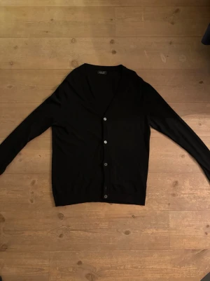 Svart kofta från Zara Man - Klassisk svart kofta från Zara Man med v-ringning och knappar framtill. Tillverkad i mjuk bomullsmix och har långa ärmar. Perfekt för lager på lager och enkel att matcha med olika outfits.