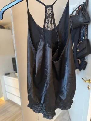 Svart linnetopp med spetsdetalj - Säljer en svart linnetopp i glansigt material med smala axelband och snygg spetsdetalj i ryggen. Toppen har en lös passform och resår nertill som ger en ballongliknande siluett. Perfekt för dig som gillar en chic och enkel look.