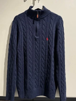 Marinblå stickad polotröja Polo Ralph Lauren - Säljer en marinblå stickad polotröja från Polo Ralph Lauren med klassiskt kabelmönster och röd broderad logga på bröstet. Tröjan har half zip med läderdetalj och ribbade muddar vid ärmslut och nederkant. Perfekt för kyliga dagar.