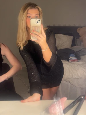 Svart glittrig kortklänning med öppen rygg - Supersnygg svart kortklänning med glitter och lång ärm. Klänningen har en djup öppen rygg med knytband i nacken och sitter tight. Perfekt för dig som vill sticka ut med en glittrig look. Materialet känns stretchigt och bekvämt.