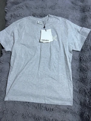 Grå t-shirt från Kenzo i bomull - Snygg grå t-shirt från Kenzo med vit logga tryckt på bröstet. Klassisk rund halsringning och korta ärmar. Tillverkad i mjuk bomull för skön känsla och enkel stil. Perfekt till jeans eller shorts för en clean look.