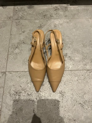 Beige pumps med spetsig tå från Sappun - Snygga beige pumps från Sappun med spetsig tå och smala remmar med guldfärgade spännen. Inköpt i Seoul, aldrig använda. Skorna har låg klack och är gjorda i ett slätt syntetmaterial som ger en clean och stilren look. Perfekta för dig som gillar minimalistisk stil.