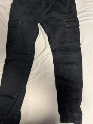 Svarta cargopants från Saligia XS - Svarta cargopants från Saligia i storlek XS. Byxorna har stora fickor med kardborrestängning på sidorna och resår vid bensluten. Tillverkade i ett mjukt bomullsmaterial och har en klassisk cargomodell med normal passform.