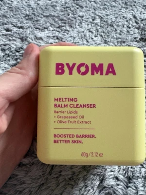 BYOMA Melting Balm Cleanser 60g - Säljer en BYOMA Melting Balm Cleanser i en ljusgul fyrkantig burk med lila text. Innehåller 60g rengöringsbalm med barrier lipids, druvkärneolja och olivfruktextrakt. Perfekt för att ta bort smink och boosta hudbarriären.