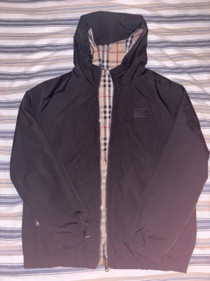 Svart vindjacka från Burberry - Säljer en svart/beige reversibel vindjacka från Burberry med klassiskt rutigt foder och huva. Jackan har dragkedja framtill, elastiska muddar och diskret Burberry-logga på bröstet. Perfekt för dig som vill ha en stilren jacka med snygga detaljer.