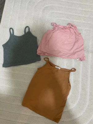 Tre linnen i olika färger - Säljer tre linnen med smala axelband. Ett ribbat mörkgrått linne, ett ljusrosa linne med volangkant upptill och ett brunt linne i mjuk bomull. Perfekta för sommaren och lätta att matcha med jeans eller kjol.