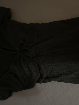Svart långklänning med snörning - Säljer en svart långklänning i mjukt, strukturerat tyg. Klänningen har korta ärmar och snygg snörningsdetalj framtill vid bysten. Passformen är normal och tyget känns lätt och luftigt, perfekt för varmare dagar.