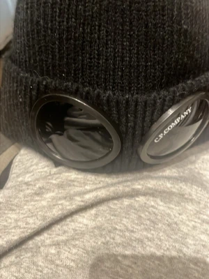 Svart mössa med goggles från C.P. Company - Säljer en svart ribbstickad mössa från C.P. Company med ikoniska goggles framtill. Mössan är tillverkad i 100% bomull och har en cool, urban look som sticker ut. Perfekt accessoar för dig som gillar streetwear och vill ha något unikt.