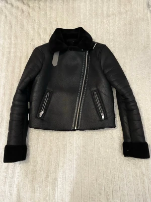 Svart pälsfodrad skinnjacka storlek xs/s - Säljer en svart pälsfodrad skinnjacka med schyssta detaljer som sned dragkedja, två fickor med dragkedjor och spännen i sidorna. Jackan har en bred krage och uppvikta ärmslut med päls. Perfekt för dig som vill ha en edgy och varm look.