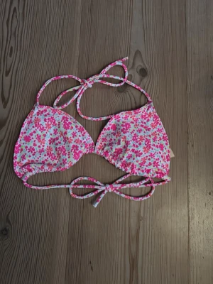 Blommig bikiniöverdel i rosa och vitt - Söt trekantsbikini med blommigt mönster i rosa, vitt och lite grönt. Knytband i nacken och ryggen för justerbar passform. Perfekt för strandhäng och soliga dagar.