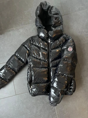 Svart glansig pufferjacka från Colmar - Säljer en svart, glansig pufferjacka från Colmar med huva och dragkedja framtill. 