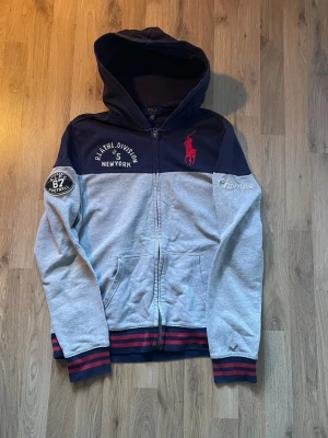 Polo Ralph Lauren Hoodie - Snygg och bekväm zip-hoodie från Polo Ralph Lauren i marinblått och grått med röda detaljer. Broderad logga framtill samt patches på ärm och bröst ger en sportig och exklusiv  Fråga gärna! Snabb leverans!