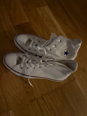 Vita Converse All Star high tops - Klassiska vita Converse All Star high tops med röd och blå rand runt sulan. Skorna har snörning, rund tå och är tillverkade i canvas. Converse-logga på sidan och 'All Star'-märke bak på hälen. Perfekta till jeans eller shorts för en clean och tidlös look.