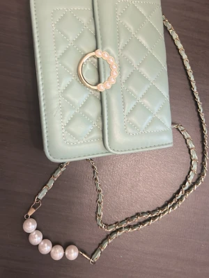 Mintgrön quiltad axelväska med pärldetaljer - Snygg mintgrön axelväska i quiltad design med guldfärgad kedja och pärlor på både låset och axelbandet. Väskan har en elegant ringdetalj framtill och är perfekt för dig som vill ha något unikt och trendigt. Materialet känns mjukt och lyxigt.