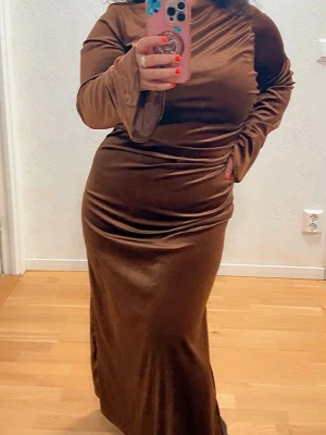 Brun långärmad sammetsklänning - Säljer en elegant brun långklänning i mjuk sammet med långa ärmar och figurnära passform. Klänningen har en enkel, stilren design med rund halsringning och lätt utsvängda ärmslut. Perfekt för dig som vill ha en chic och bekväm look.