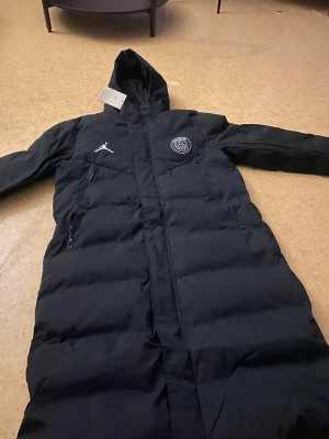 Svart lång pufferjacka PSG x Jordan - Svart lång pufferjacka med huva från Jordan x PSG. Jackan har vita loggor på bröstet, två sidofickor med dragkedja och quiltad design. Perfekt för kalla dagar och har en clean, sportig vibe. STORLEK M. DET ÄR INTE SOM DEM ANDRA JACKORNA