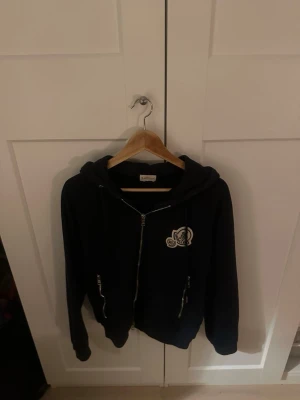 Svart cardigan jacka från Moncler - Svart cardigan jacka från Moncler med huva, dragkedja och broderad logga på bröstet. Jackan har snörning vid huvan och två fickor med dragkedja. Materialet är mjukt och känns riktigt skönt mot huden, perfekt för chill dagar.