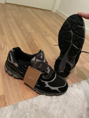 Svarta och grå Asics sneakers - Säljer ett par snygga Asics sneakers i svart och grått med coola detaljer. Ovandelen är i mesh och syntet med tydliga loggor på sidan. Skorna har snörning och en rejäl, stötdämpande sula som passar perfekt till streetwear eller träning.