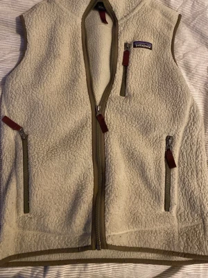 Beige fleeceväst från Patagonia - Säljer en beige fleeceväst från Patagonia med dragkedja framtill och tre fickor med dragkedja. Västen har bruna detaljer längs kanterna och är perfekt till alla möjliga olika klädstilar säljer denna nu pågrund av att den inte används 