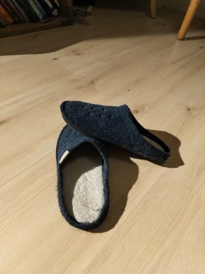 Mörkblå tofflor från Crocs i ull - Mörkblå tofflor från Crocs med ovandel i filtad ull och innersula i ljus ullmix. Yttersulan är i slitstark syntet med mönstrad undersida för bra grepp. Klassisk slip-in modell med rund tå och enkel design, perfekta för att chilla hemma.