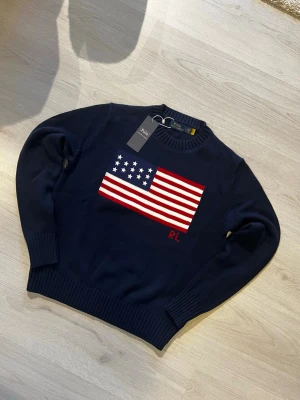 Mörkblå stickad tröja Polo Ralph Lauren - Mörkblå stickad tröja från Polo Ralph Lauren med amerikansk flagga i rött, vitt och blått på bröstet samt RL-broderi. Klassisk rund halsringning och ribbade muddar vid ärmslut och nederkant. Perfekt för dig som gillar preppy stil.