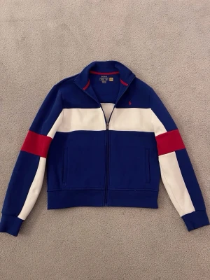 Snygg Ralph lauren zip tröja - Säljer en snygg Ralph lauren zip tröja i mörkblå, röd och vit färg. Tröjan är i nytt skick och ändats använd ett fåtal gånger. Perfekt för dig som vill ha en snygg mysig tröja för vardags bruk eller vid finare tillfällen.