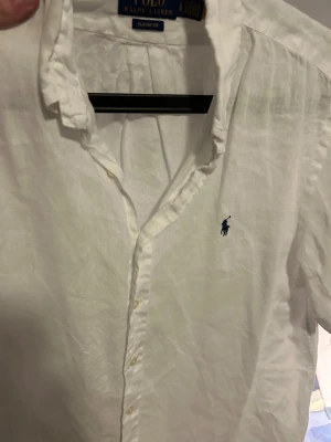 Vit linneskjorta från Polo Ralph Lauren - Vit klassisk skjorta från Polo Ralph Lauren i storlek L med korta armar. Tillverkad i linne med klassisk passform och knappar framtill. Har den ikoniska blå Polo-loggan broderad på bröstet och en snygg krage. Perfekt för en clean och fräsch look.