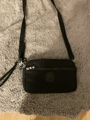 Svart axelväska med dragkedja - Säljer en svart axelväska från Kipling med justerbar rem och flera fack med dragkedja. Väskan är i ett lätt syntetmaterial och har en rund Kipling-logga framtill. Perfekt storlek för att bära det viktigaste och enkel att matcha till olika outfits.