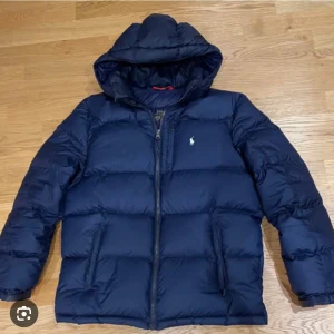Marinblå pufferjacka från Polo Ralph Lauren - Säljer en marinblå pufferjacka från Polo Ralph Lauren med huva och dragkedja. Jackan har den klassiska loggan på bröstet och två sidofickor. Perfekt för kalla dagar och riktigt snygg till vardagslooken.