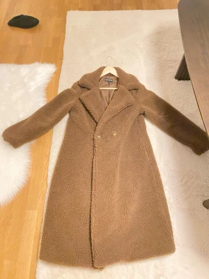 Brun teddyjacka från EllyBazar - Säljer en lång, brun teddyjacka från EllyBazar med bred krage och dubbelknäppning framtill. Jackan har ett mjukt, fluffigt material och är helfodrad på insidan. Perfekt för dig som vill ha en mysig och trendig look under kyliga dagar.