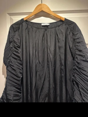 Svart långklänning från Stockh LM - Säljer en svart långklänning från Stockh LM med unika plisserade långärmar och rund halsringning. Klänningen har en loose passform och är tillverkad i ett lätt, glansigt material som ger en snygg drapering. Perfekt för dig som gillar statement-ärmar och stilren design.