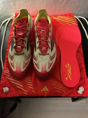 Adidas F50 fotbollsskor röd/vit - Säljer ett par Adidas F50 fotbollsskor med snygg design i rött, vitt och gult. Skorna har snörning, platt sula med dobbar och svarta Adidas-ränder på sidan. Ovandelen är i syntetmaterial och innersulan är röd med F50-tryck. Perfekt för dig som vill sticka ut på planen. Box samt det andra medföljer. Hör gärna av er för frågor!
