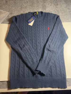 Mörkblå kabelstickad tröja Polo Ralph Lauren - Kabelstickad mörkblå tröja från Polo Ralph Lauren med klassisk röd logga broderad på bröstet. Tröjan har rund halsringning, ribbade muddar och är långärmad. Perfekt för kyliga dagar och enkel att styla till jeans eller chinos.